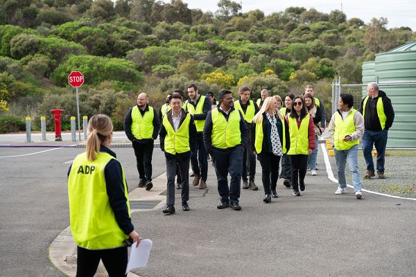A group of people, male and female in a mixture of business clothing and casual wear are walking on a wide road inside a water treatment facility. They are all wearing high visibility vests and are walking toward a woman at the front of the photo who has her back to camera. 
